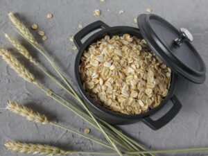Flat lay of rolled oats in a black pot surrounded by wheat stalks on a textured surface.